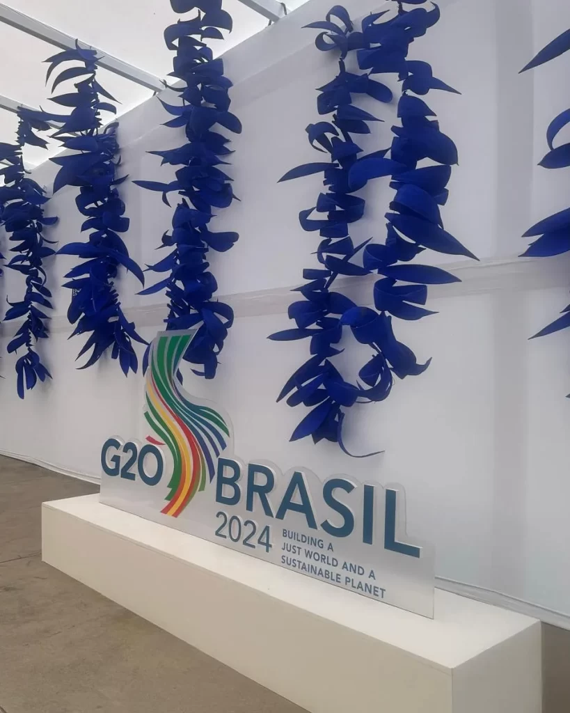 Visão lateral de diversas tiras de Côco azul penduradas em cachos na atrás do totem do G20 Brasil Rio Summit na entrada da Sala de Imprensa.